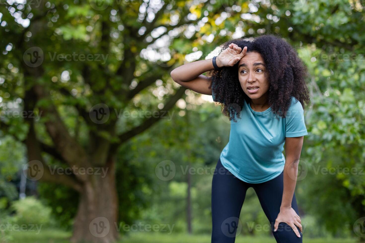 African American woman in athletic wear takes a break during outdoor exercise in park. She appears determined yet fatigued, displaying motivation and perseverance against lush green background under sunny summer skies. photo