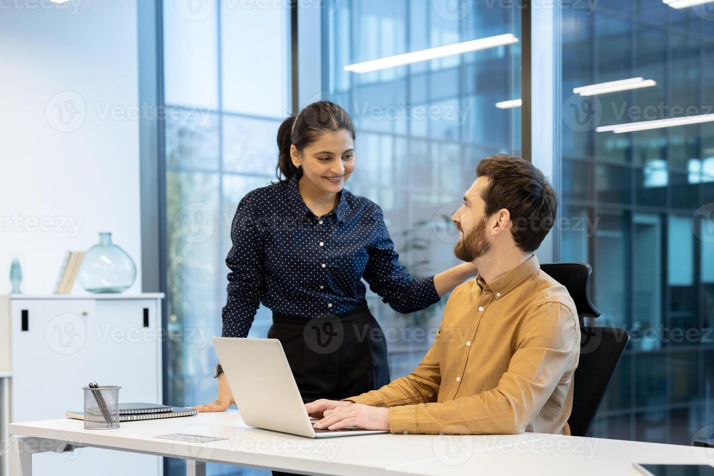 A woman smiles while placing her hand on a man's shoulder as he works on a laptop in a bright office setting. photo