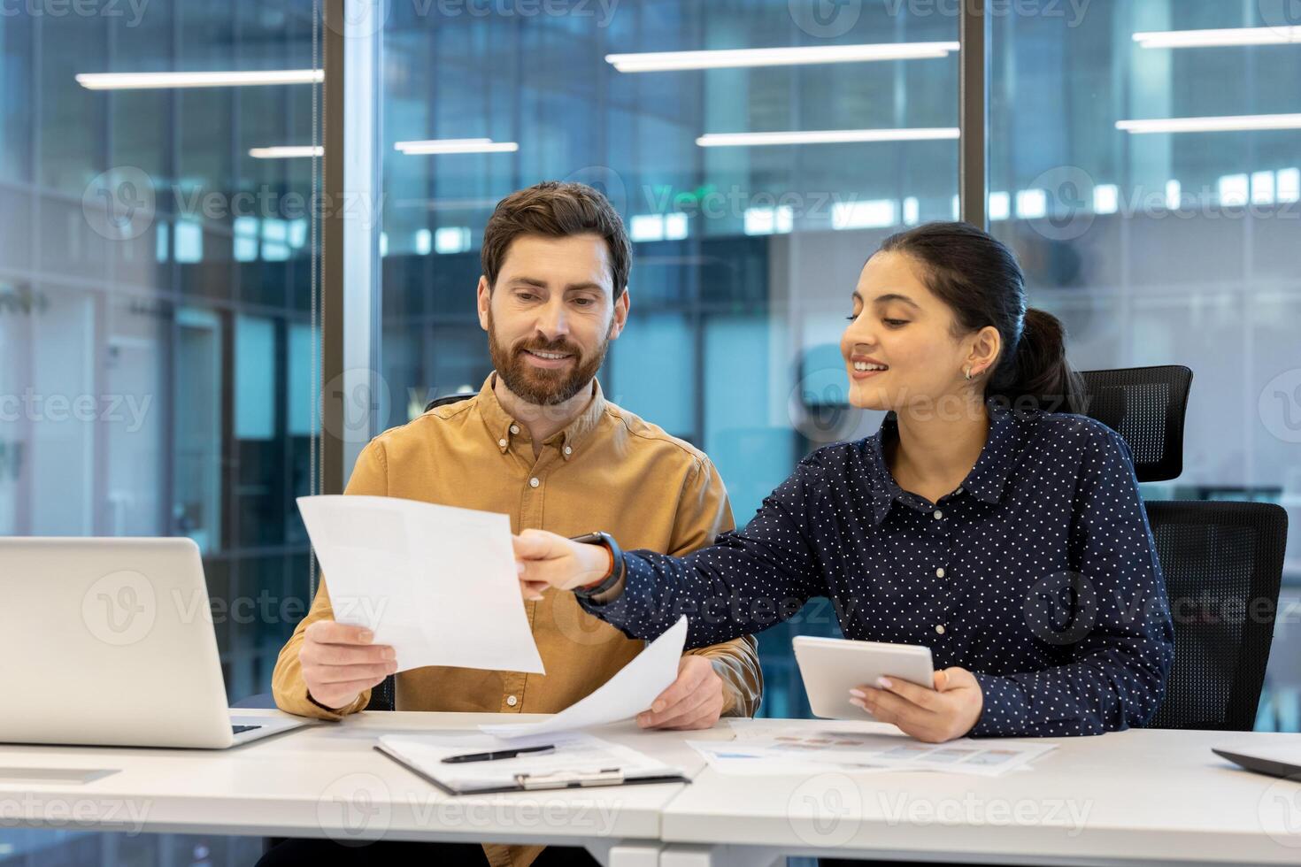 A male and female colleagues review documents at a modern office. They appear focused and engaged in a collaborative work session, discussing the papers. photo