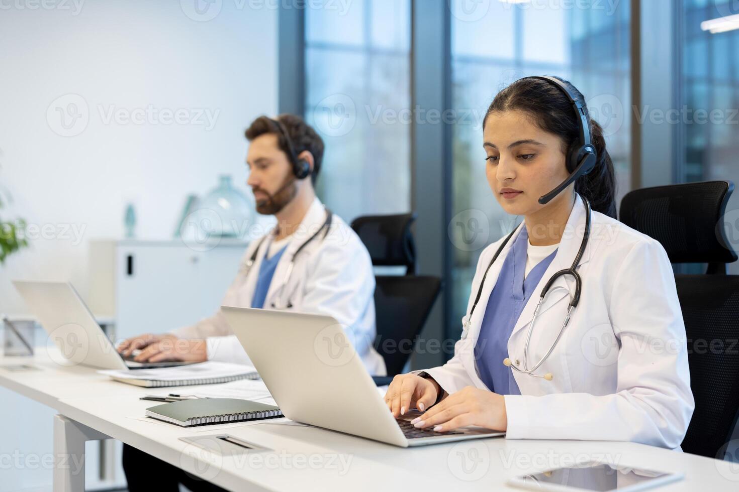 Two doctors are seen in an office setting, wearing headsets and using laptops. They may be providing remote medical consultations. photo