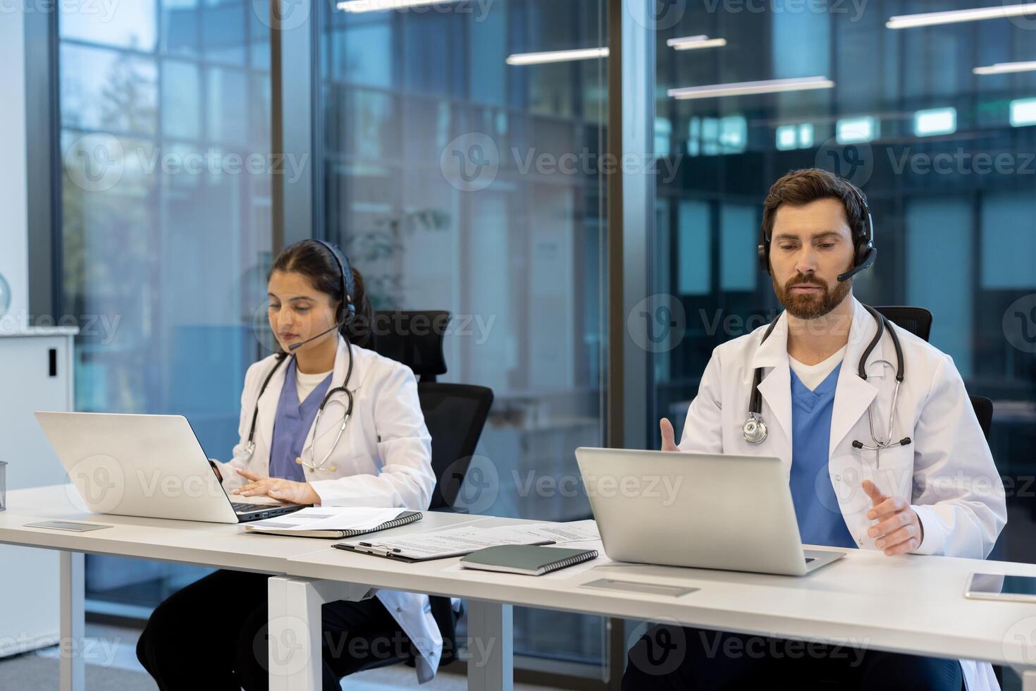 Two healthcare professionals wearing headsets assist patients remotely using laptops in a modern office setting with natural light. photo