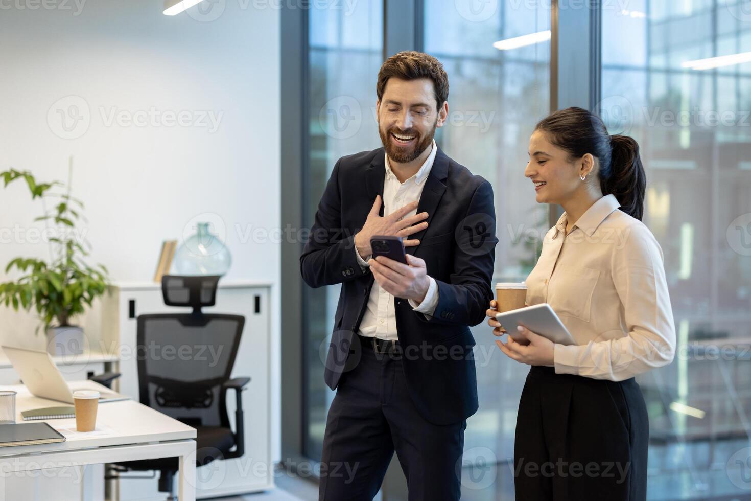 A man and a woman smile while reviewing something on a smartphone in a modern office setting, holding a coffee and tablet. photo