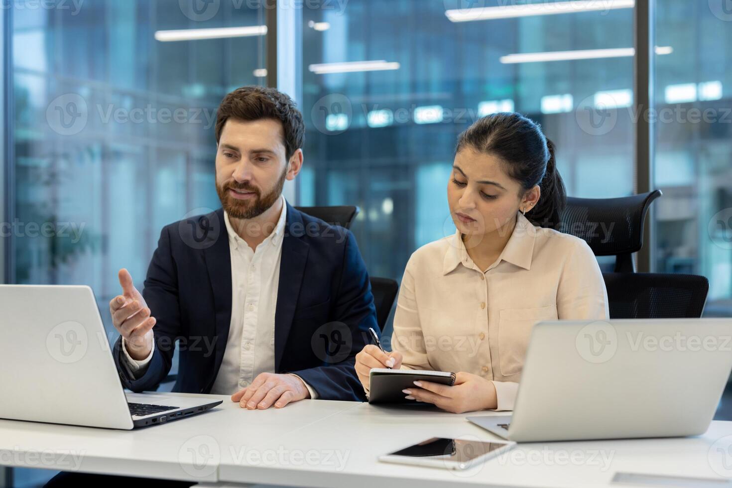 A professional man and woman collaborate in an office setting, working on laptops and taking notes during a meeting. photo