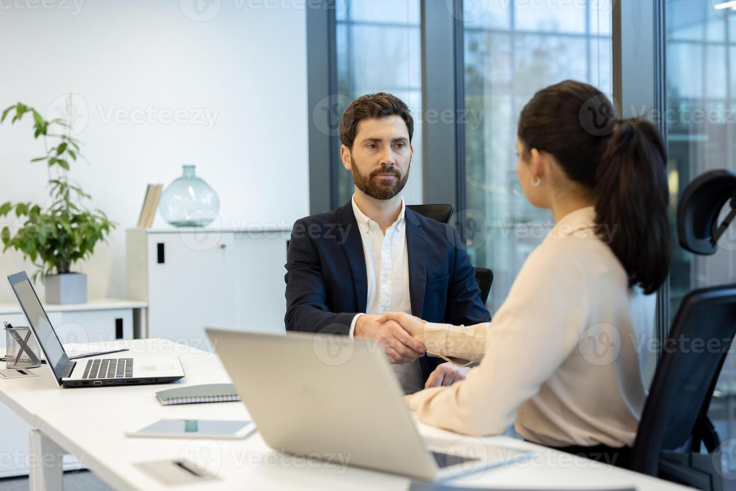 A business handshake seals a deal between two individuals seated at a modern office desk with laptops. The scene shows professional agreement and teamwork. photo
