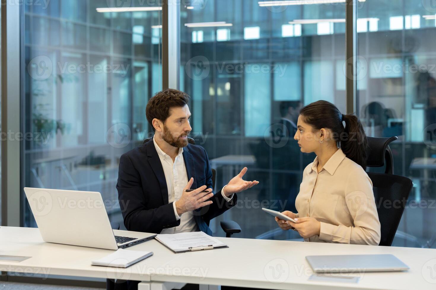 A businessman and businesswoman collaborate in an office setting, discussing using a laptop, tablet, and documents. The environment suggests a modern and professional workspace. photo