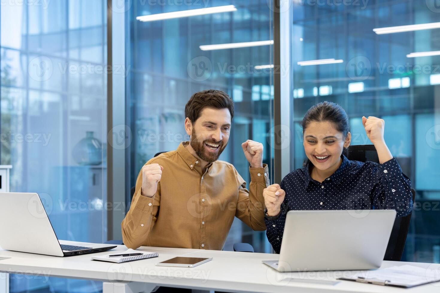 A man and a woman, thrilled by success, celebrate while working together at a modern, bright office space on their laptops. photo