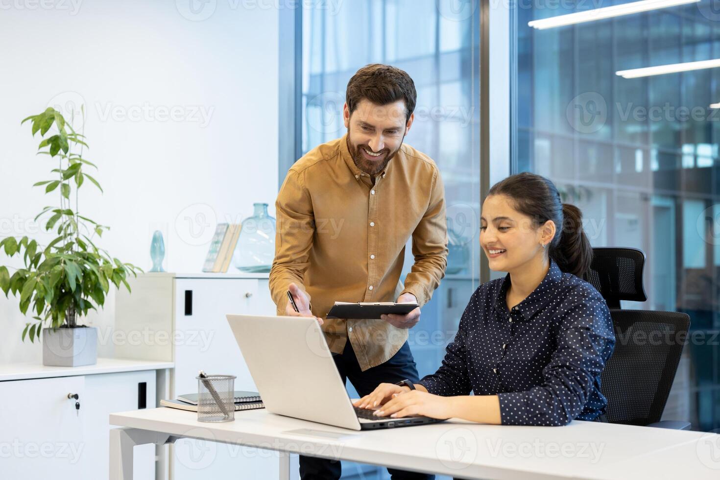 A cheerful colleague assists a female coworker as she works on a laptop in a bright modern office setting. Collaboration is key. photo