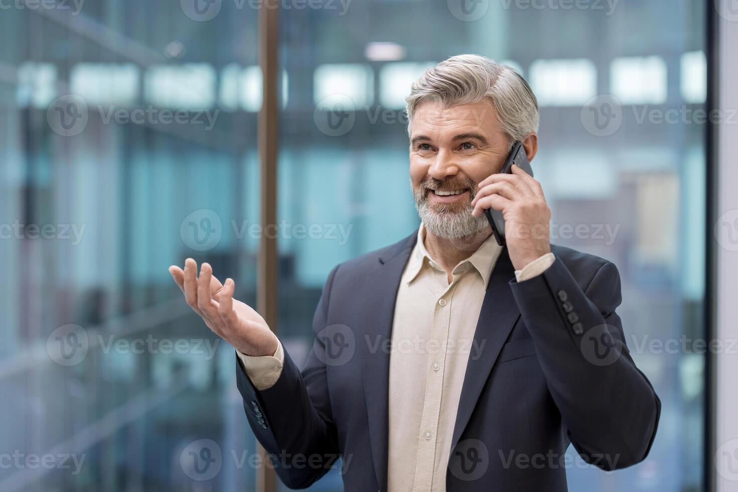 A senior executive in a suit smiles as he talks on his mobile phone in a modern office environment. He gestures with his hand. photo
