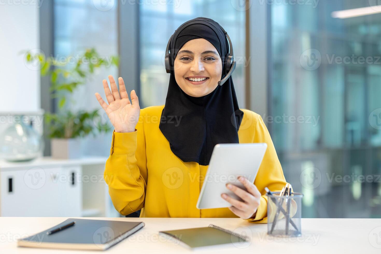 An elegant woman wearing a hijab and headset waves at the camera during a call, smiling warmly. photo