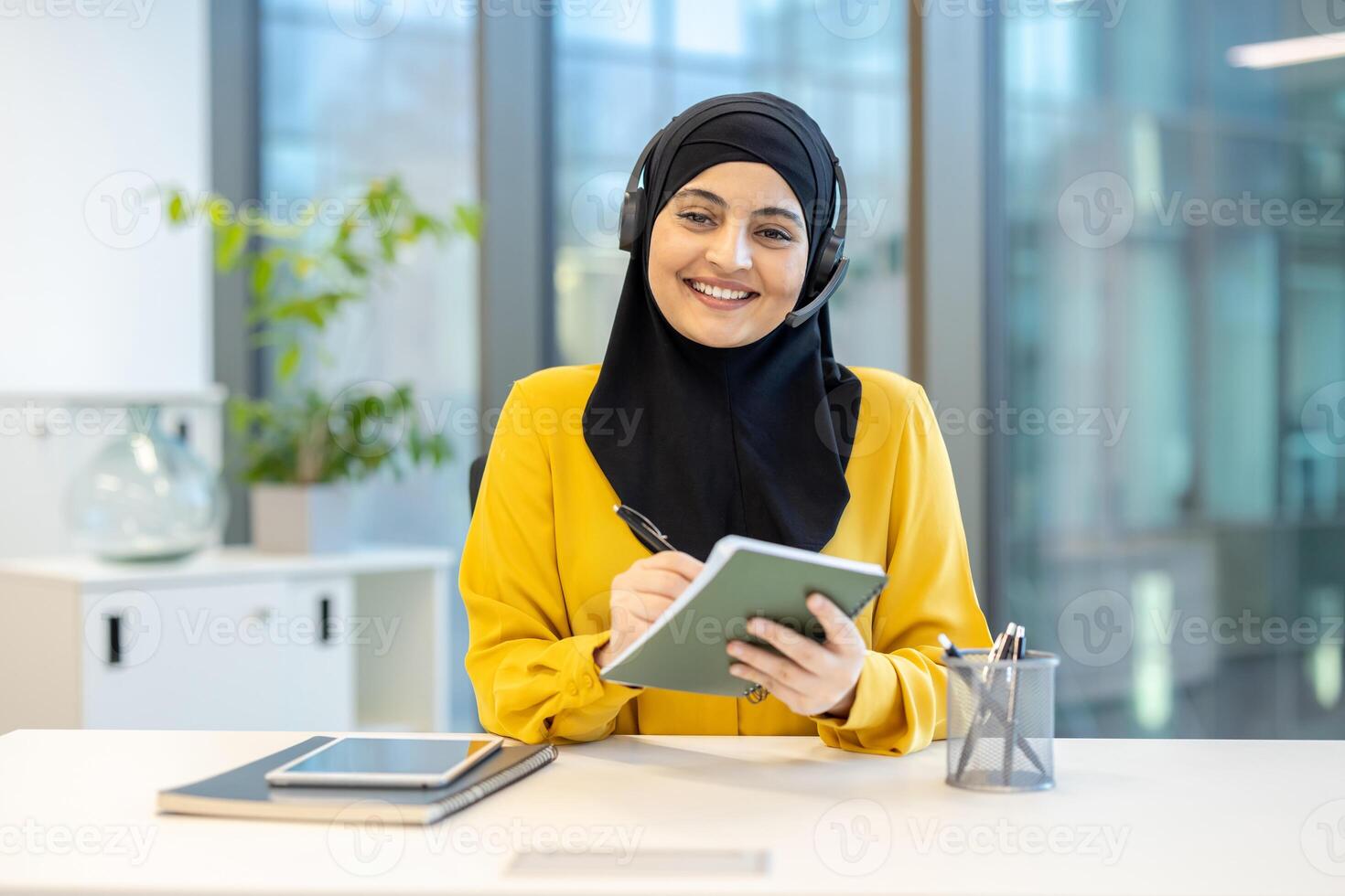 A cheerful businesswoman in a hijab wearing a headset, smiles while writing in her notebook at the office. She is working. photo