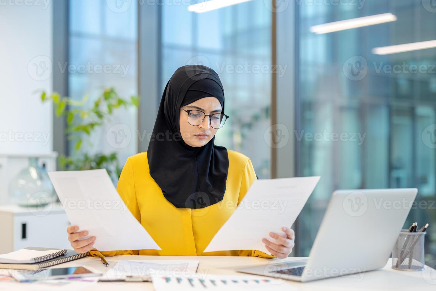 An arab woman with glasses wearing a hijab reviews documents in a modern office setting. She looks focused and concentrated at work. photo
