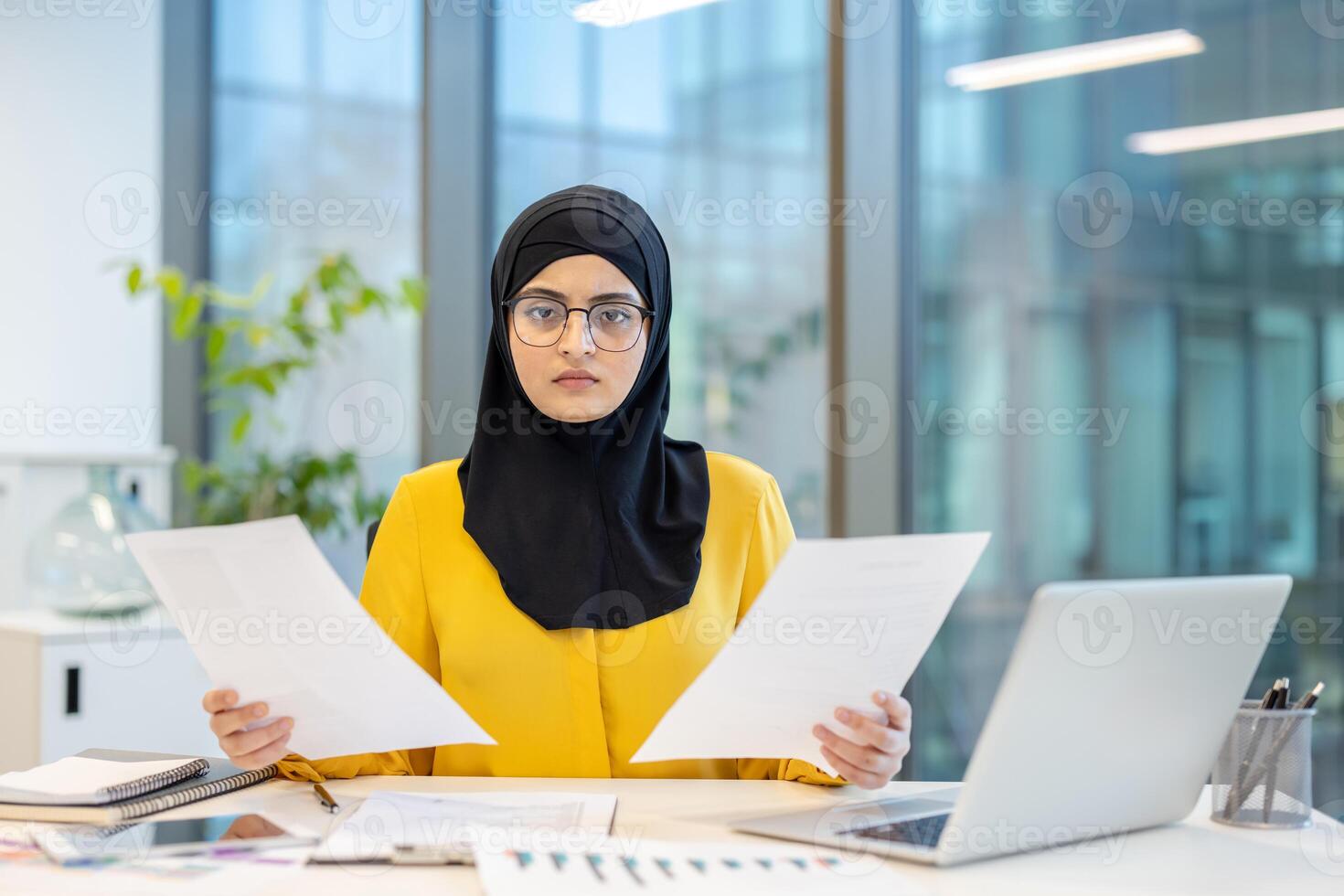 A serious-looking woman in a hijab reviews documents at her desk, using a laptop and office environment. photo