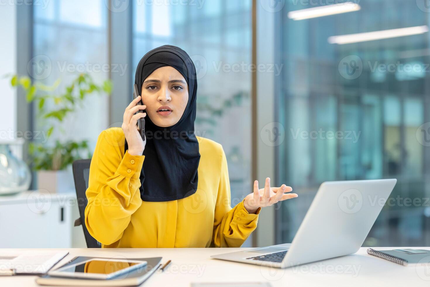A woman wearing a hijab is on the phone, looking concerned while working on her laptop in the office setting. photo
