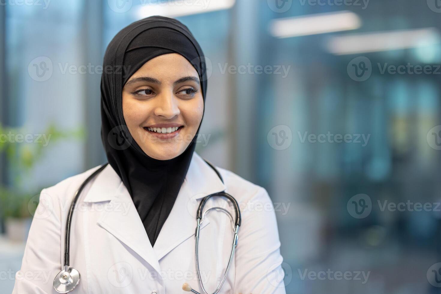 A friendly female doctor wearing a hijab and a lab coat smiles in a medical environment. Her stethoscope hangs around her neck, ready for work. photo