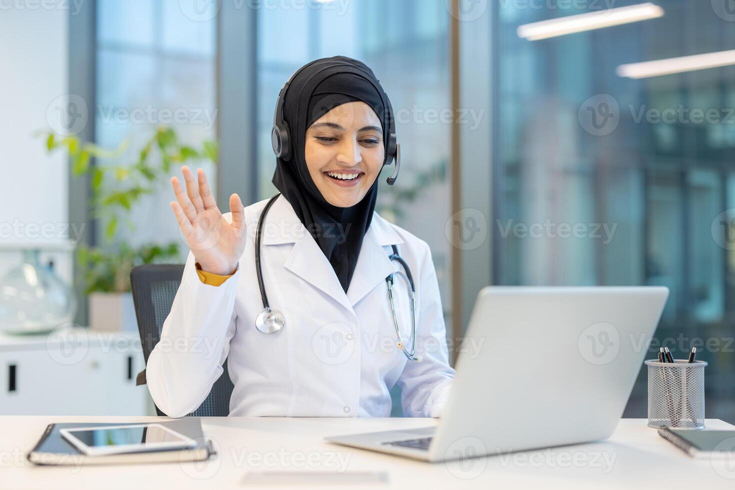 A cheerful doctor in a hijab smiles and waves during a call using a laptop, promoting telemedicine and healthcare access. photo