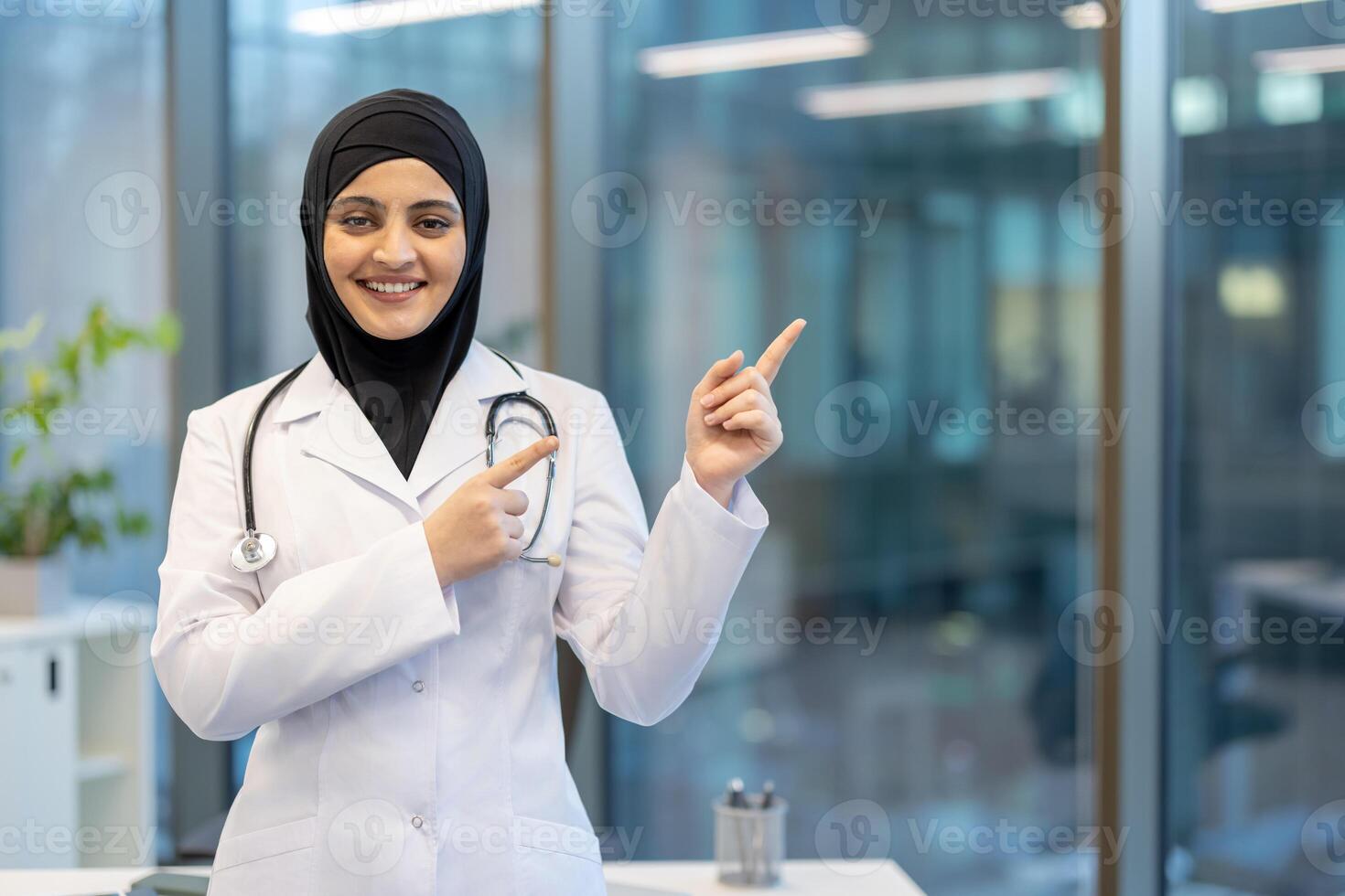 A smiling female doctor wearing a hijab and pointing towards an area in a modern medical office setting. She appears confident. photo
