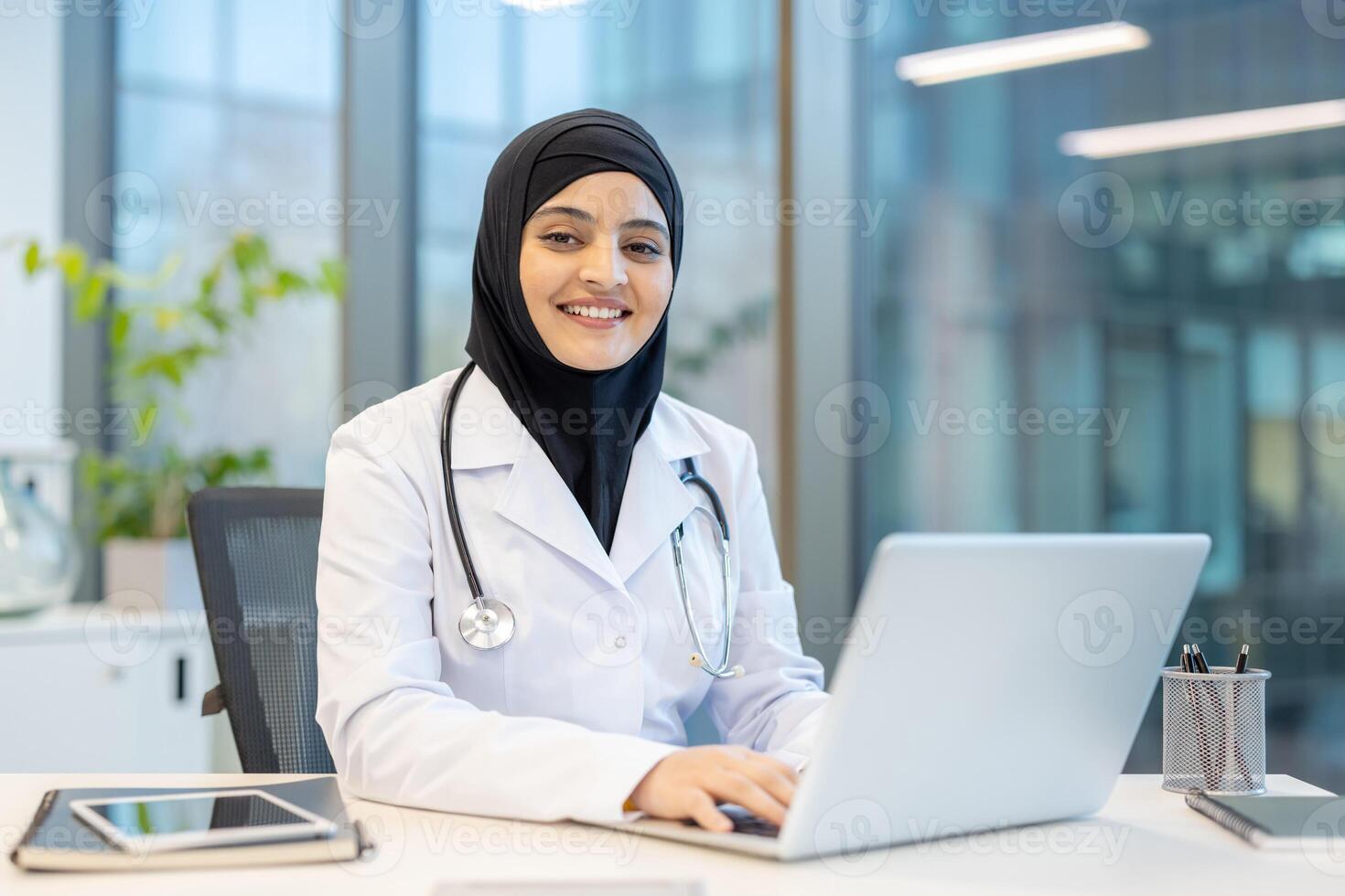 A smiling female doctor wearing a hijab sits at a desk, working on a laptop in a modern office setting, focusing on her work with a friendly demeanor. photo