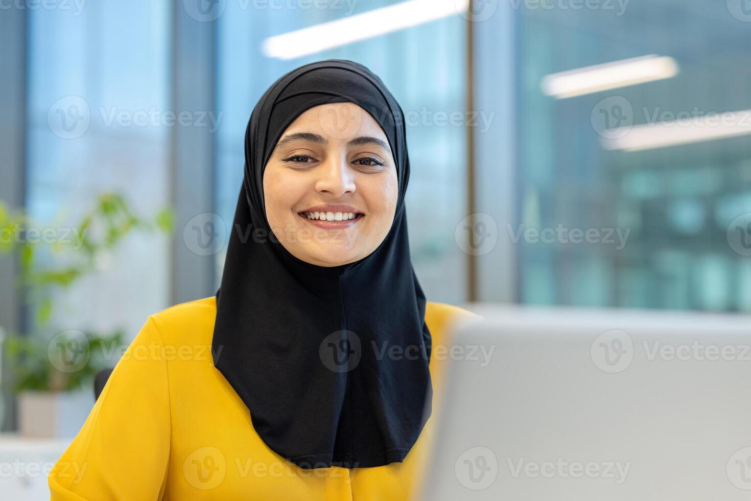 A smiling woman wearing a hijab is seen at work. She appears to be in a modern office environment with a laptop in front. photo