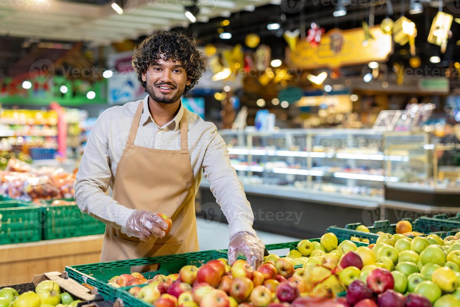 A friendly grocery store employee holds an apple while smiling at the camera, surrounded by fresh produce. photo