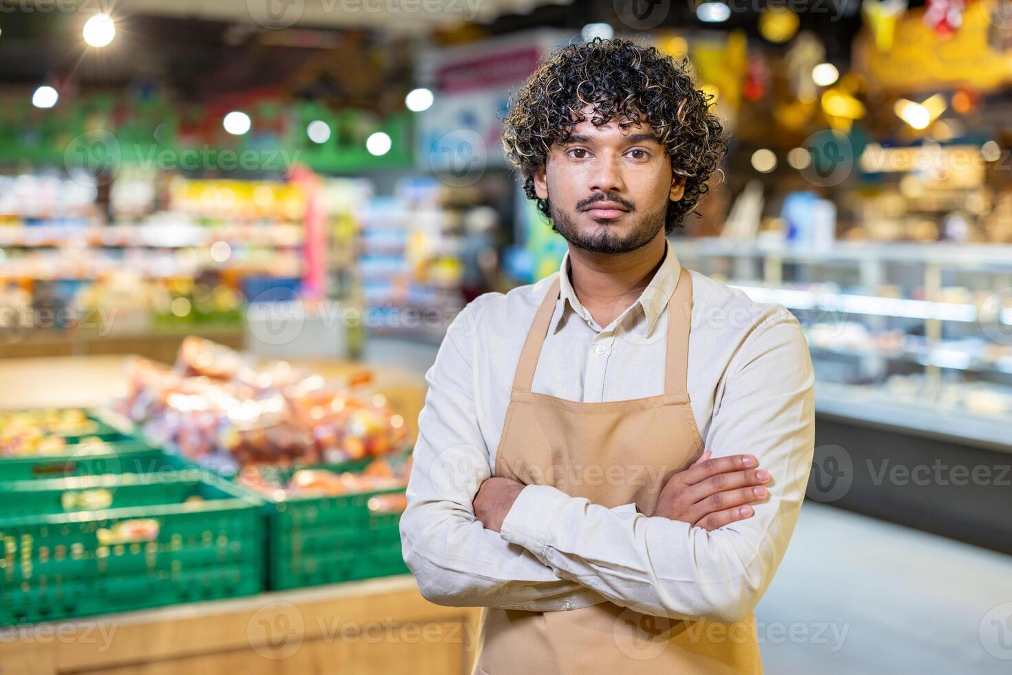 A supermarket employee, wearing an apron, poses with arms crossed while looking directly at the camera. The blurred background suggests a busy store. photo