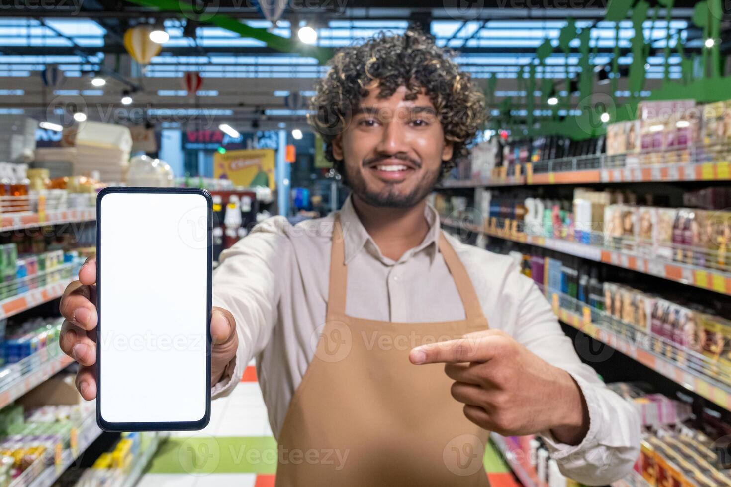 A smiling supermarket employee holds up a phone with a blank screen, inviting customers to scan, or use an app. photo