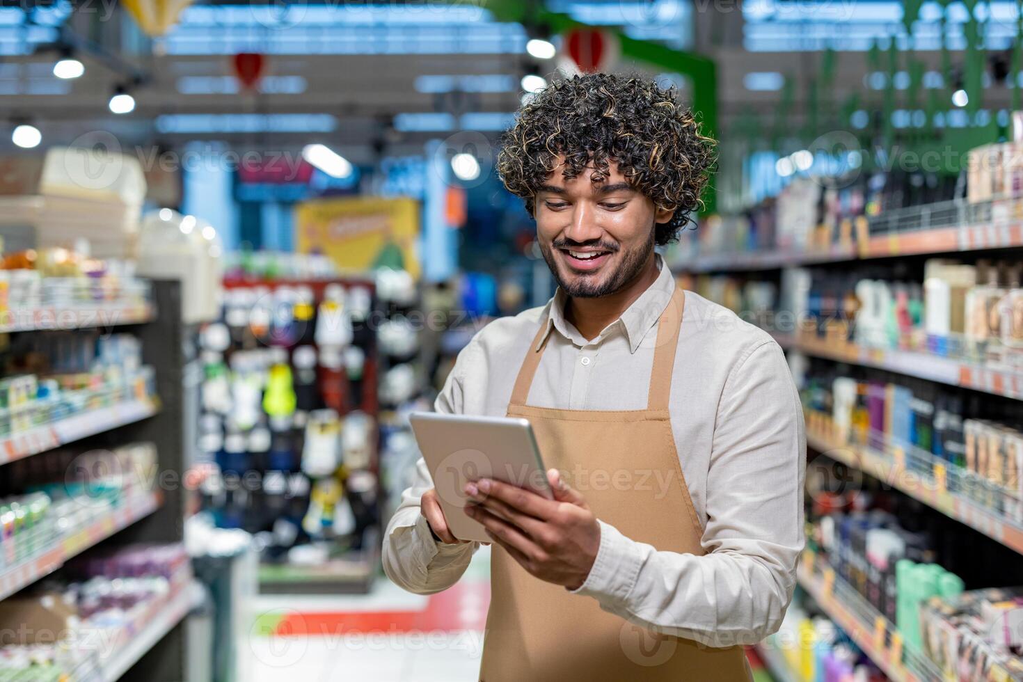 A smiling store clerk checks inventory with a tablet in a grocery store setting. Shelves filled with products surround the employee. photo