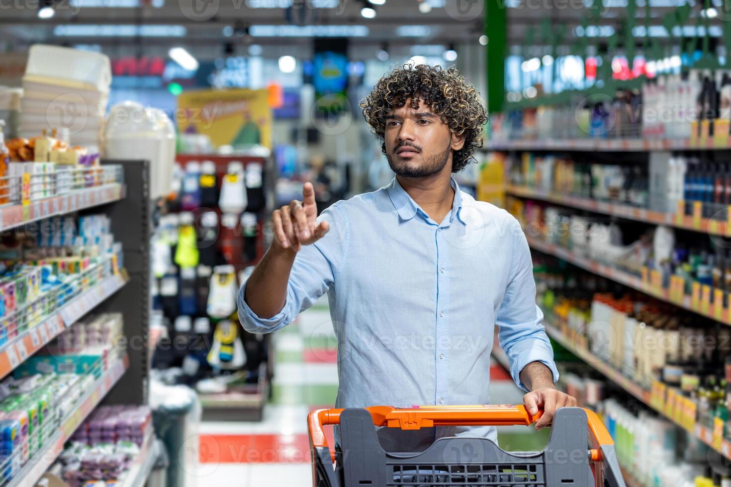 A man shops in a grocery store, extending his finger as he looks toward the shelves, pushing a shopping cart filled with items. photo