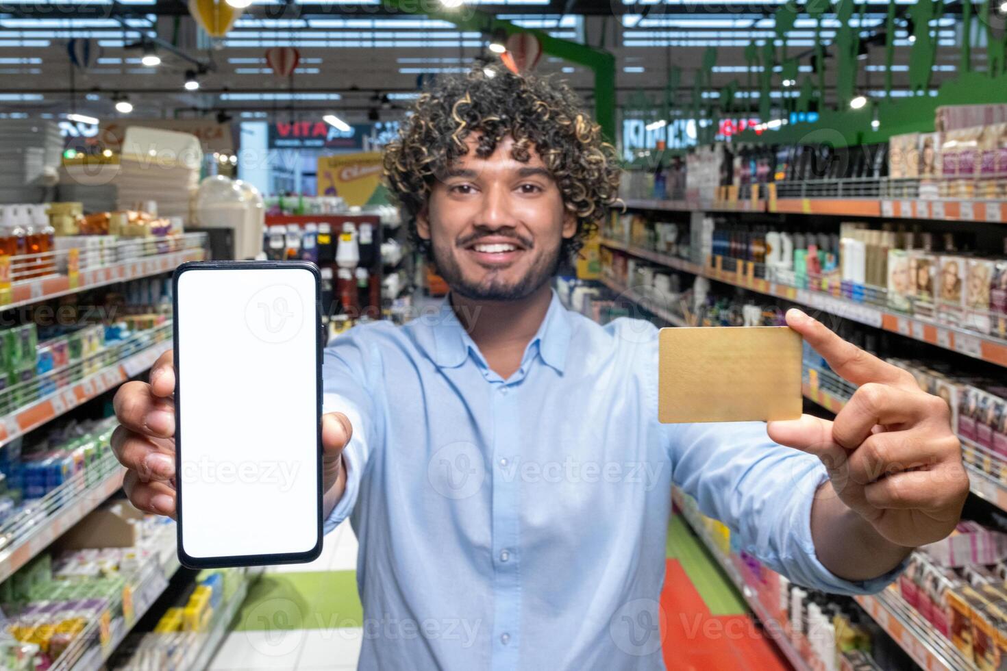 A smiling man in a grocery store holds a smartphone and a credit card, ready to shop. This scene captures convenience and payment. photo