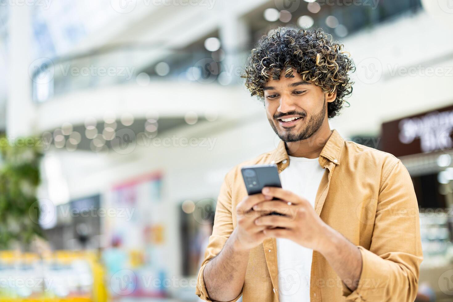 A smiling man with curly hair looks at his phone in a shopping mall, enjoying his smartphone in public. photo