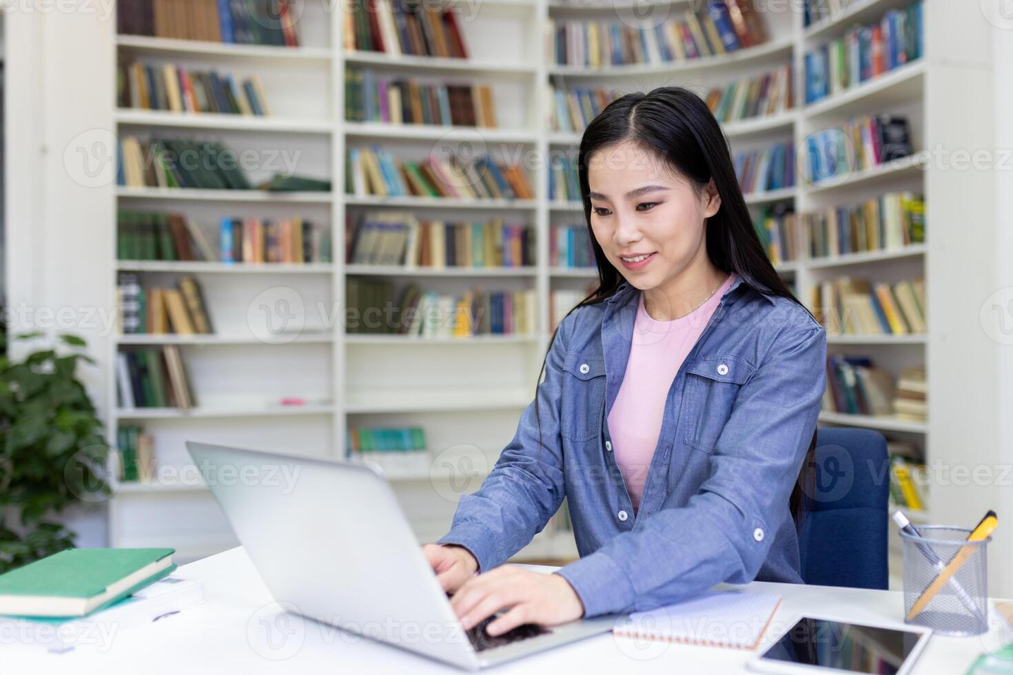 A young woman with long black hair working on her laptop in a library, books in the background. She is smiling while working on the computer. photo