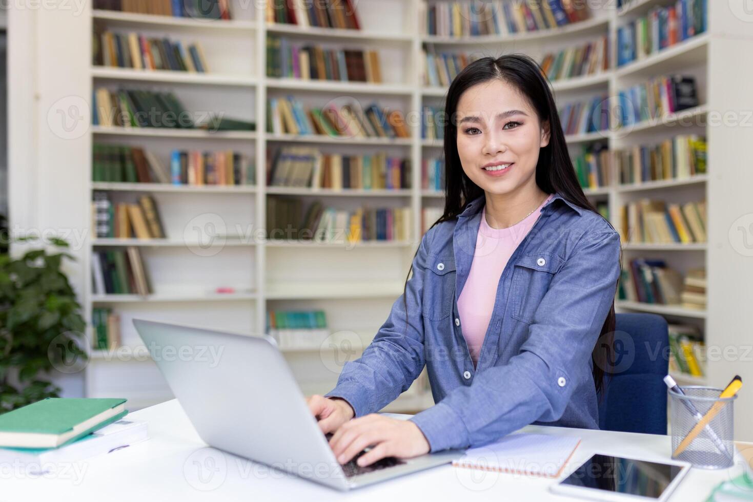 A smiling Asian woman works on her laptop at a desk in a library, surrounded by books and a focused environment. photo