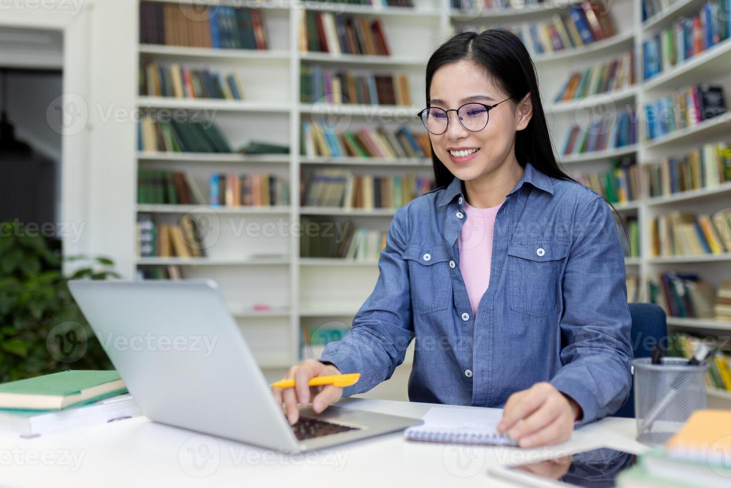 A young woman in glasses smiles as she works on a laptop in a library, surrounded by books. She appears focused and happy while engaging with her work at the desk. photo