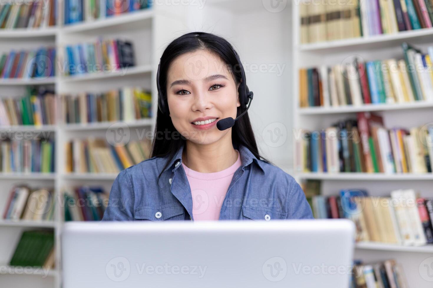 An attractive asian woman smiles while wearing a headset, working or studying remotely. The background shows bookshelves. photo