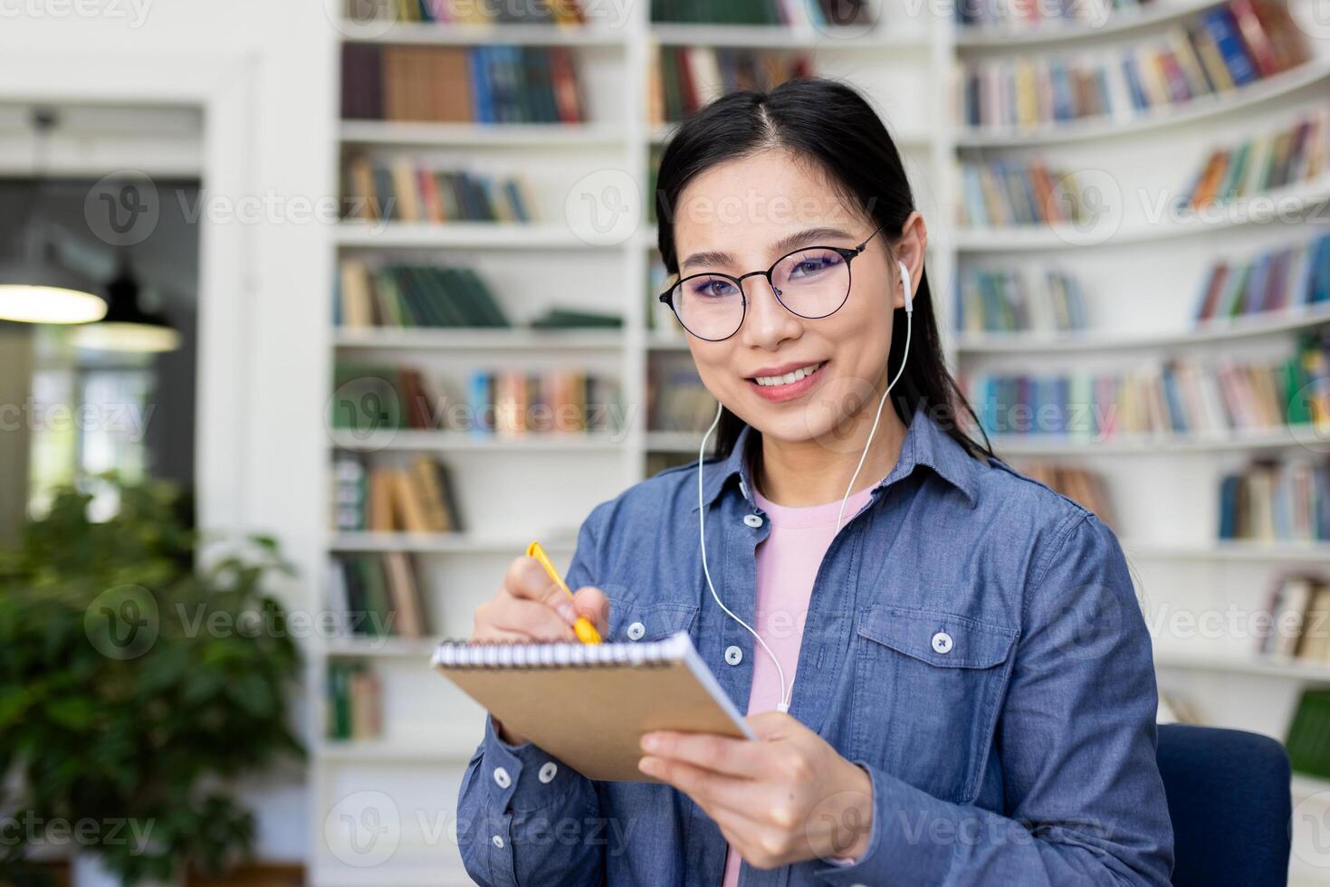 A smiling asian woman with glasses takes notes while wearing earphones, set in a library setting with a backdrop of bookshelves. photo