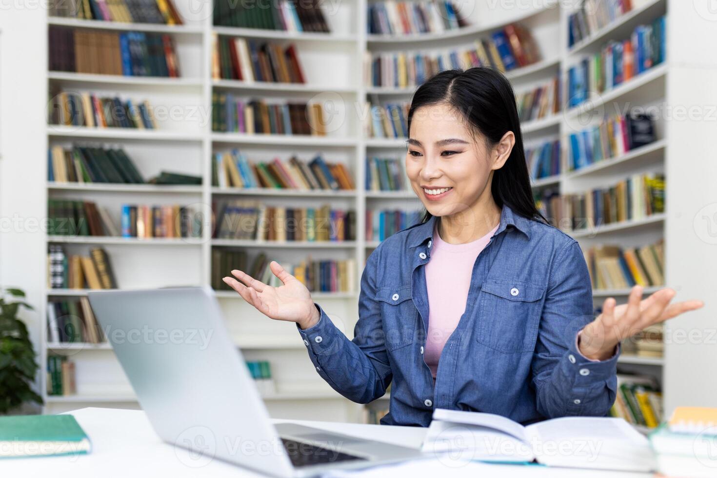 A smiling asian woman sits at a desk in a library, gesturing while looking at her laptop. Bookshelves filled with books surround her. photo