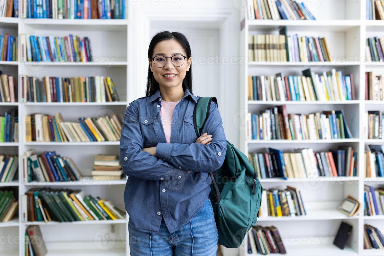 A smiling young woman stands confidently in a library, arms crossed, with a backpack, surrounded by bookshelves. She is likely a student. photo