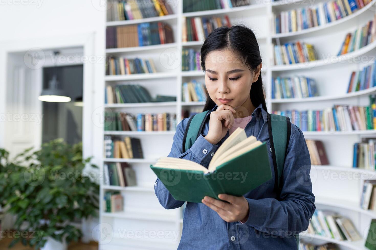 A thoughtful asian woman reads a book in a library, surrounded by bookshelves, contemplating its contents. She wears a backpack, hinting at studies. photo