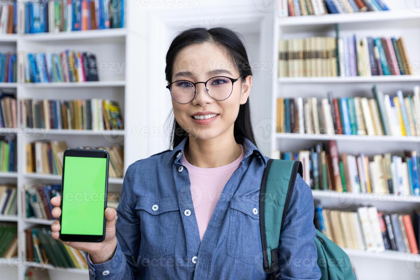 A young woman, wearing glasses and a backpack, presents a smartphone with a green screen in a library. Bookshelves line the background. photo