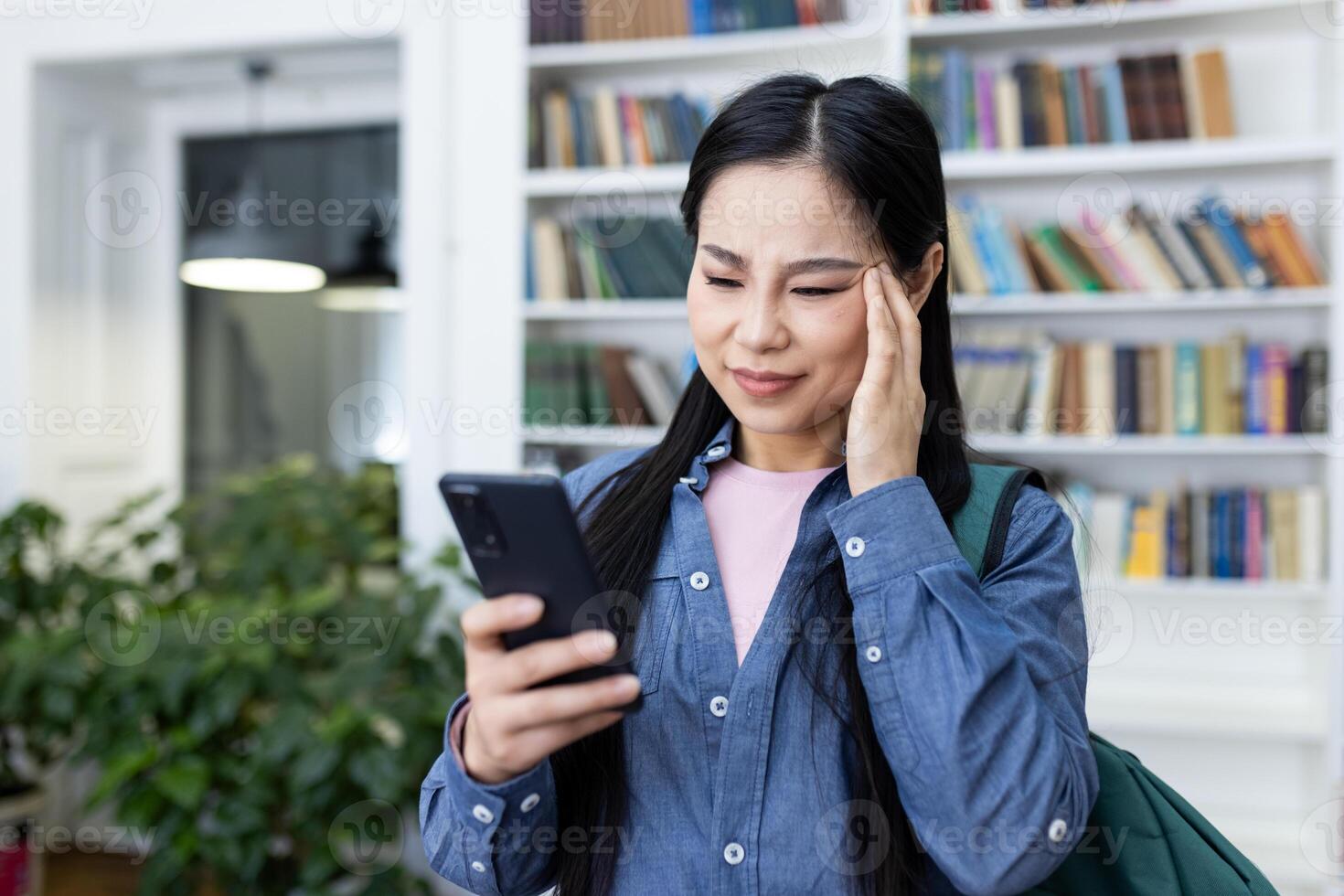 An asian woman in a library looks at her phone with a concerned expression, touching her head. A backpack is visible too. photo
