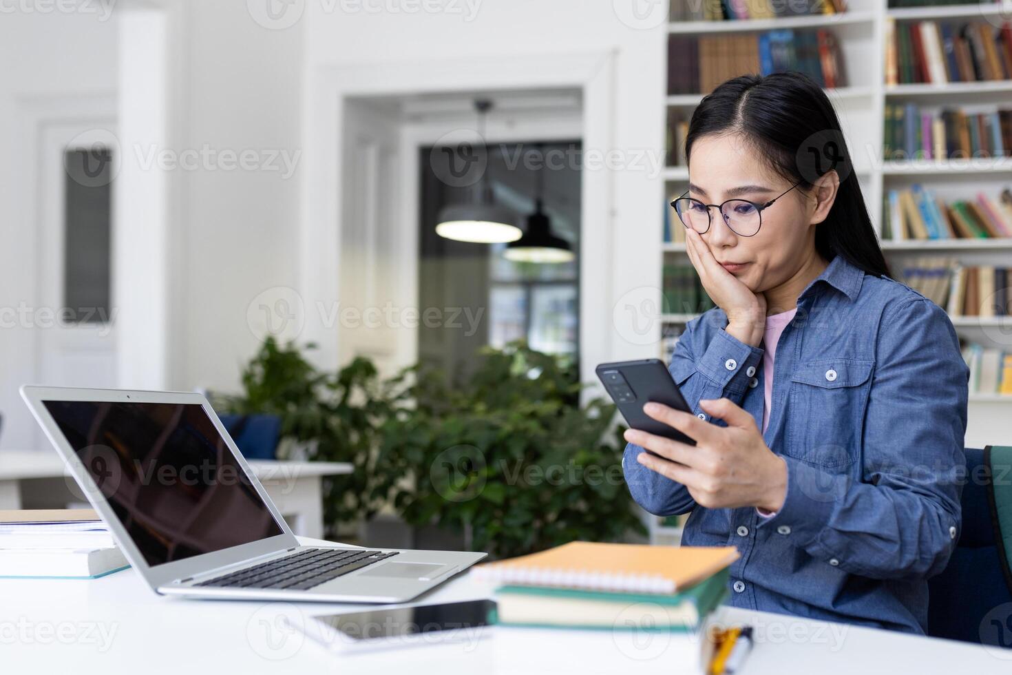 An Asian woman with glasses looks surprised while holding her phone and sitting in a library. A laptop and books are on the desk. photo