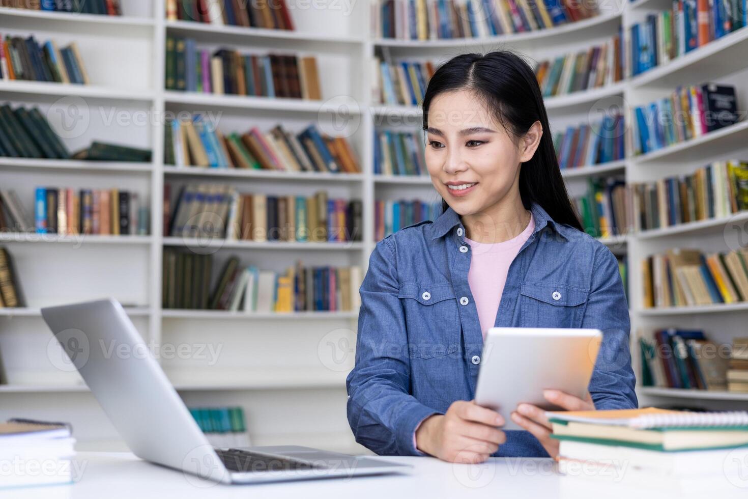 A smiling Asian woman uses a tablet while studying at a desk in a library, surrounded by books and a laptop. She seems focused. photo