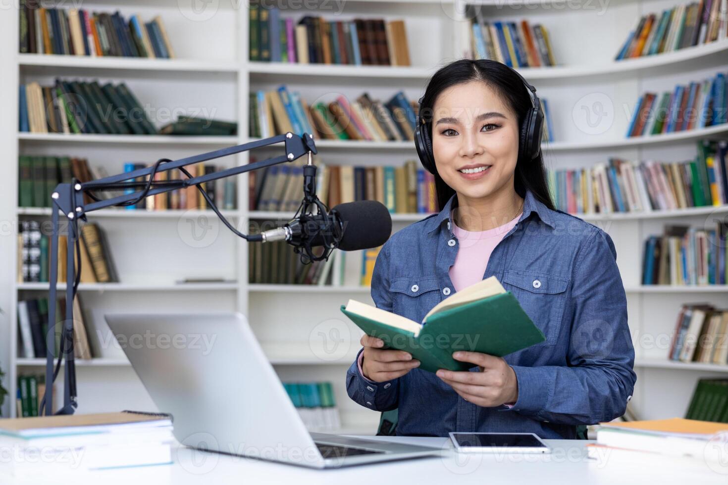 A smiling woman, wearing headphones, reads a book in a library setting while preparing to record a podcast. Bookshelves are filled with literature. photo