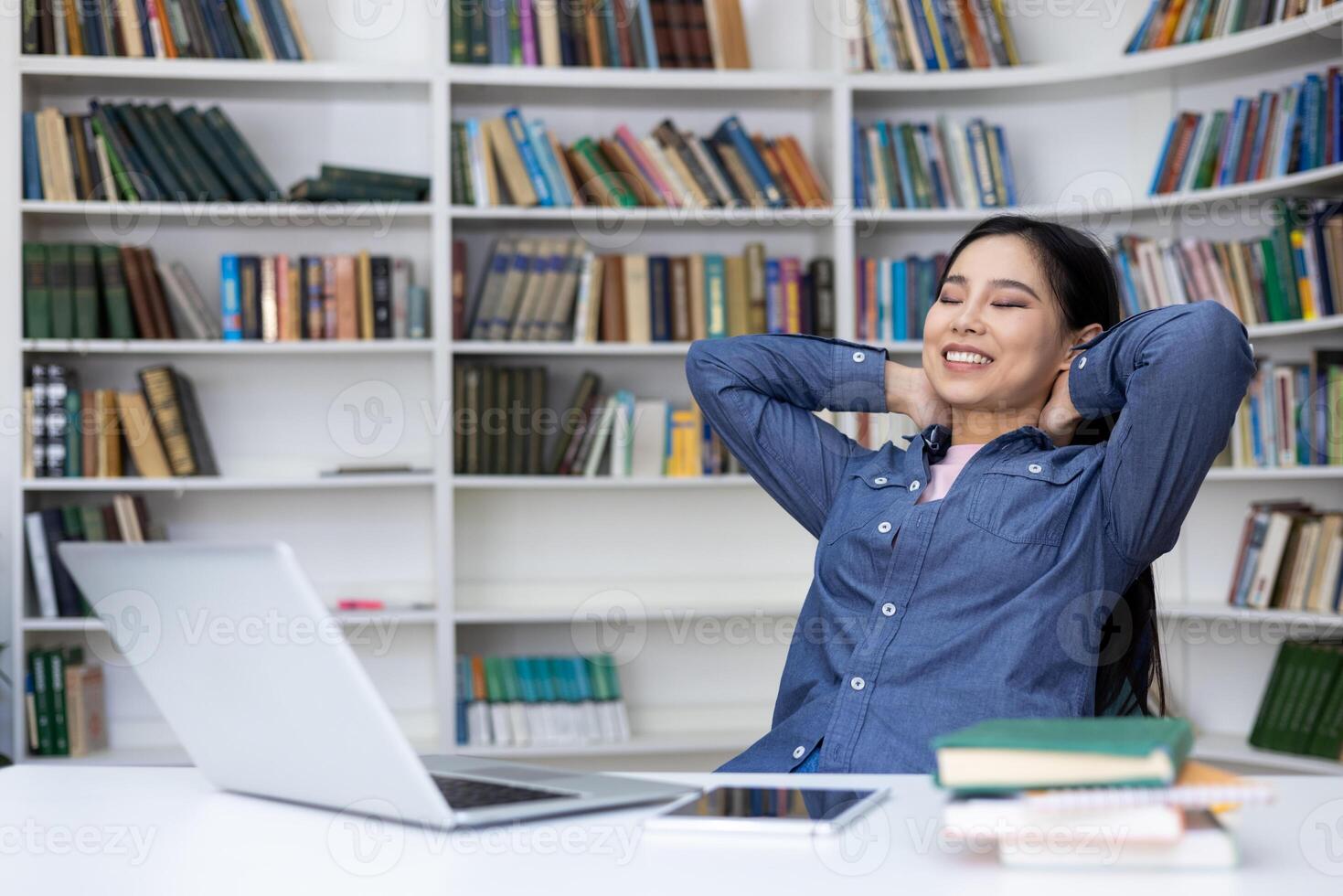 A relaxed woman smiles with her eyes closed, enjoying a break in the library. Her arms rest behind her head as she sits at her desk. Bookshelves fill the background. photo