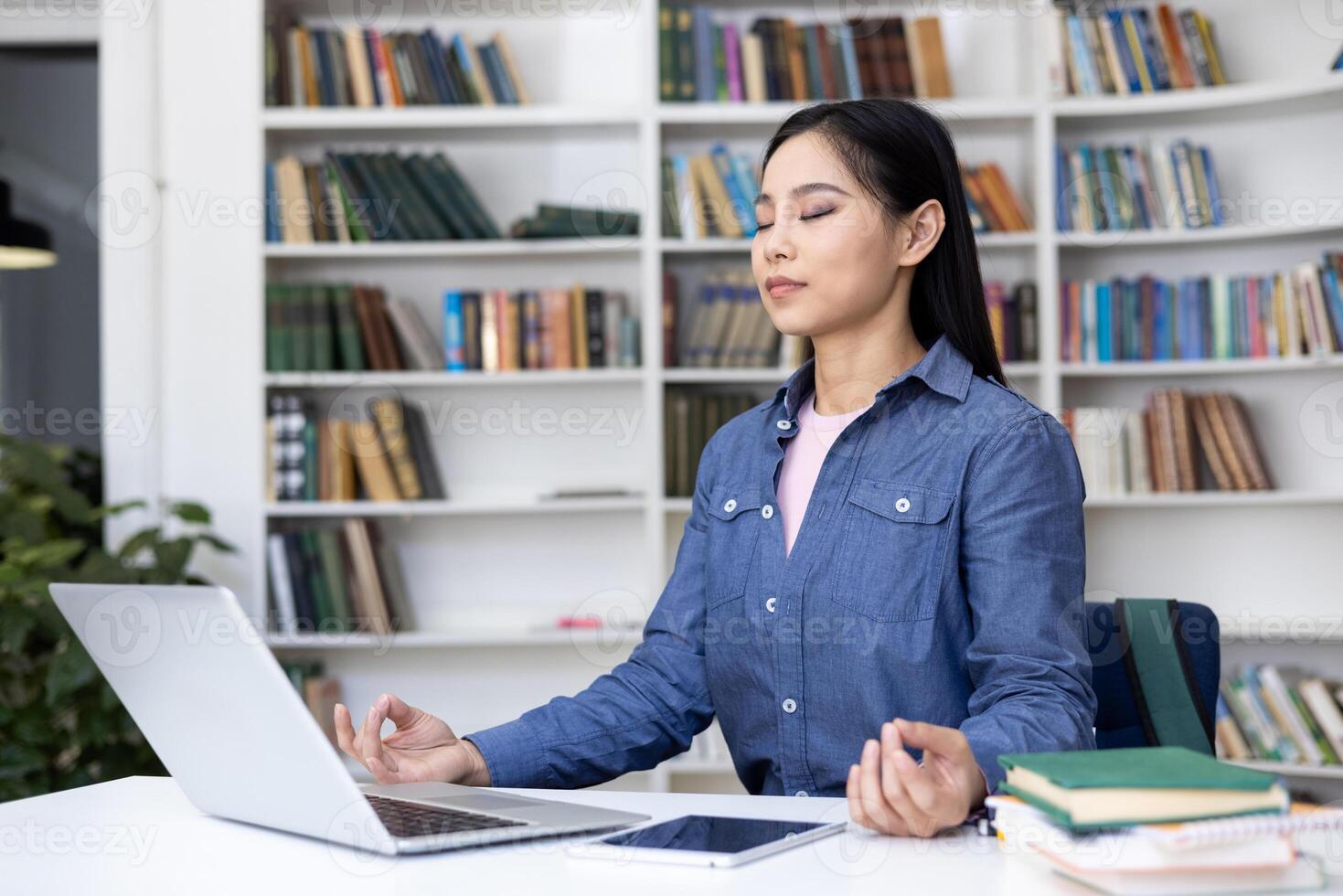 A serene Asian woman meditates in a library setting, balancing her work and well-being with mindfulness. A laptop and tablet lay nearby. photo