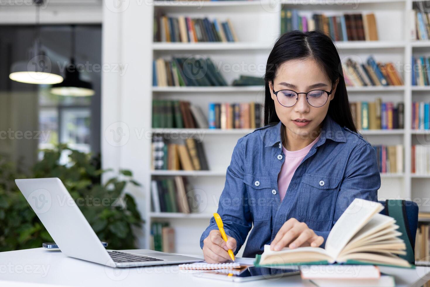 A young woman with glasses studies in a library, using books, a laptop, and taking notes. It captures the essence of learning and research. photo