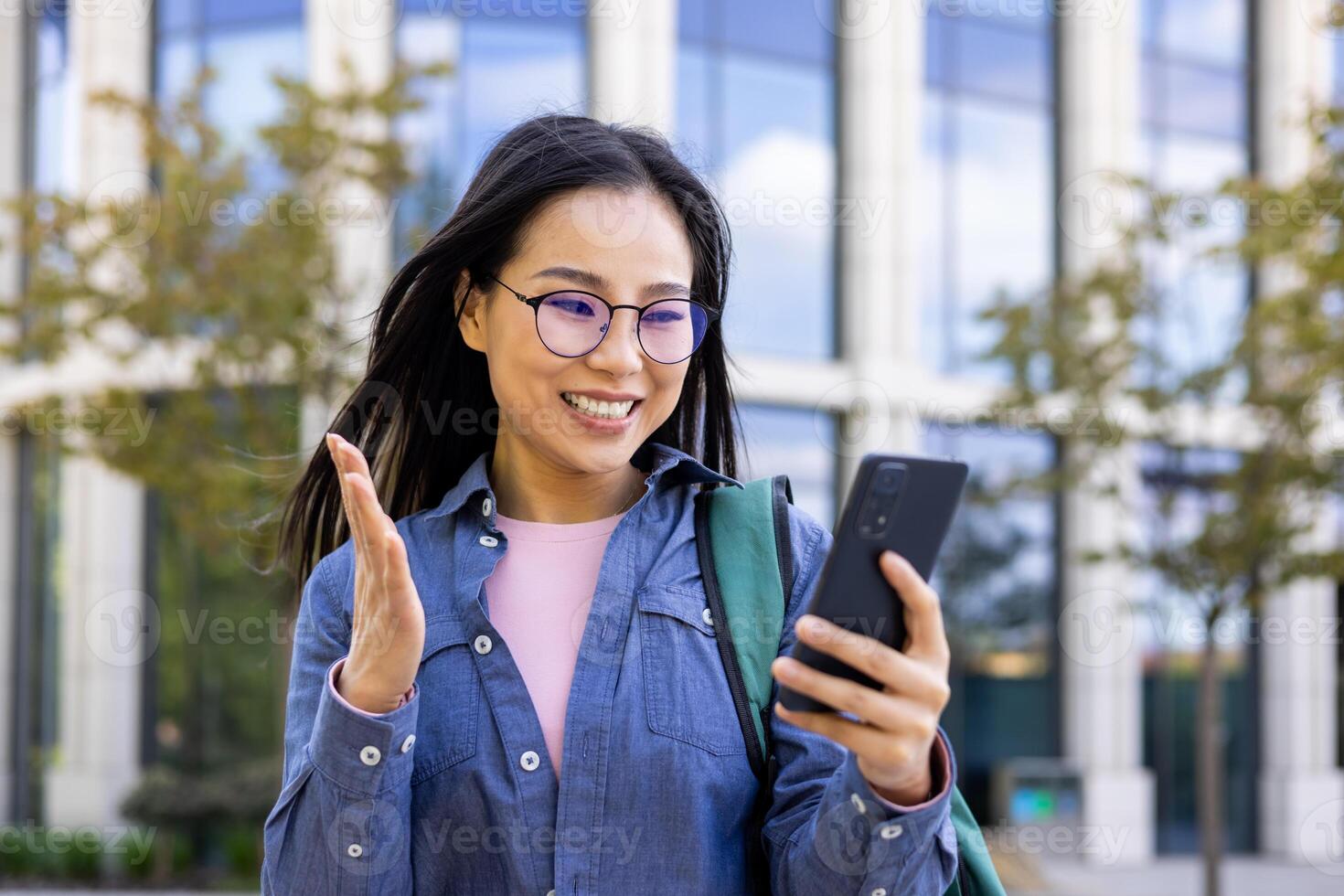 A cheerful asian woman is waving while looking at her phone. She wears eyeglasses and smiles, set against a modern building. photo