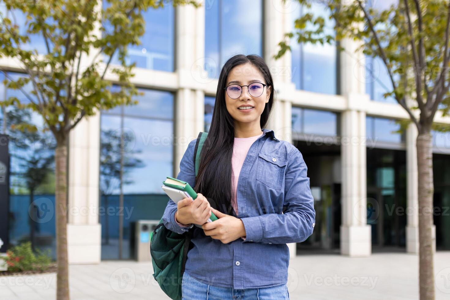A smiling asian student, wearing glasses, standing in front of a university building with books in hand, carrying a backpack. photo
