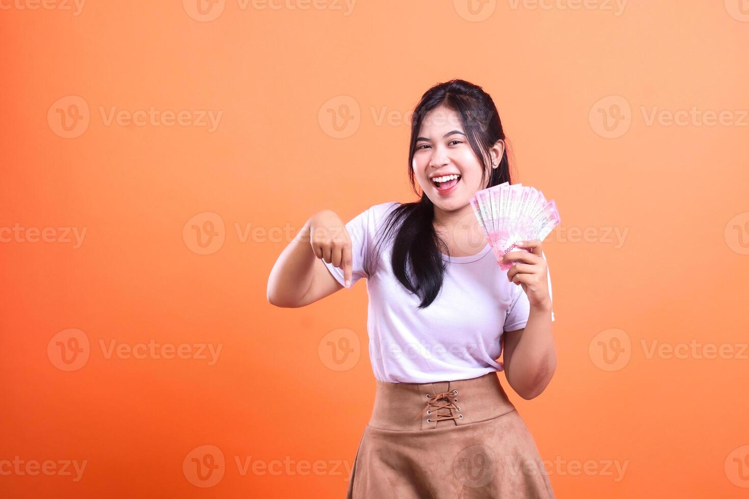 A smiling woman holds Indonesian rupiah banknotes with orange background looking at camera photo