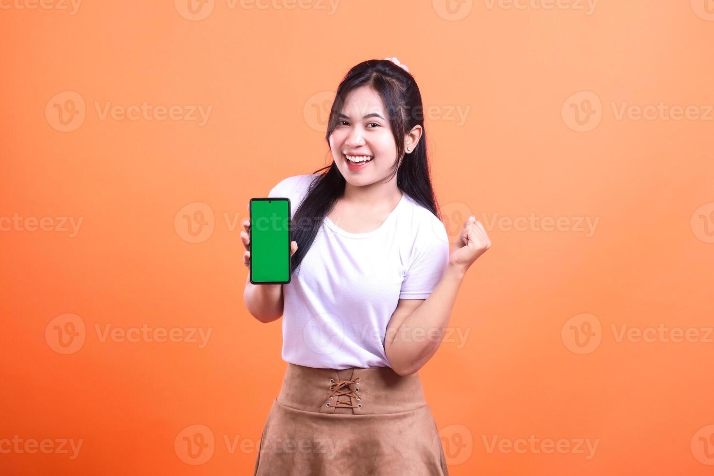 Woman in white shirt and brown skirt showcasing smartphone with green screen against an orange background photo