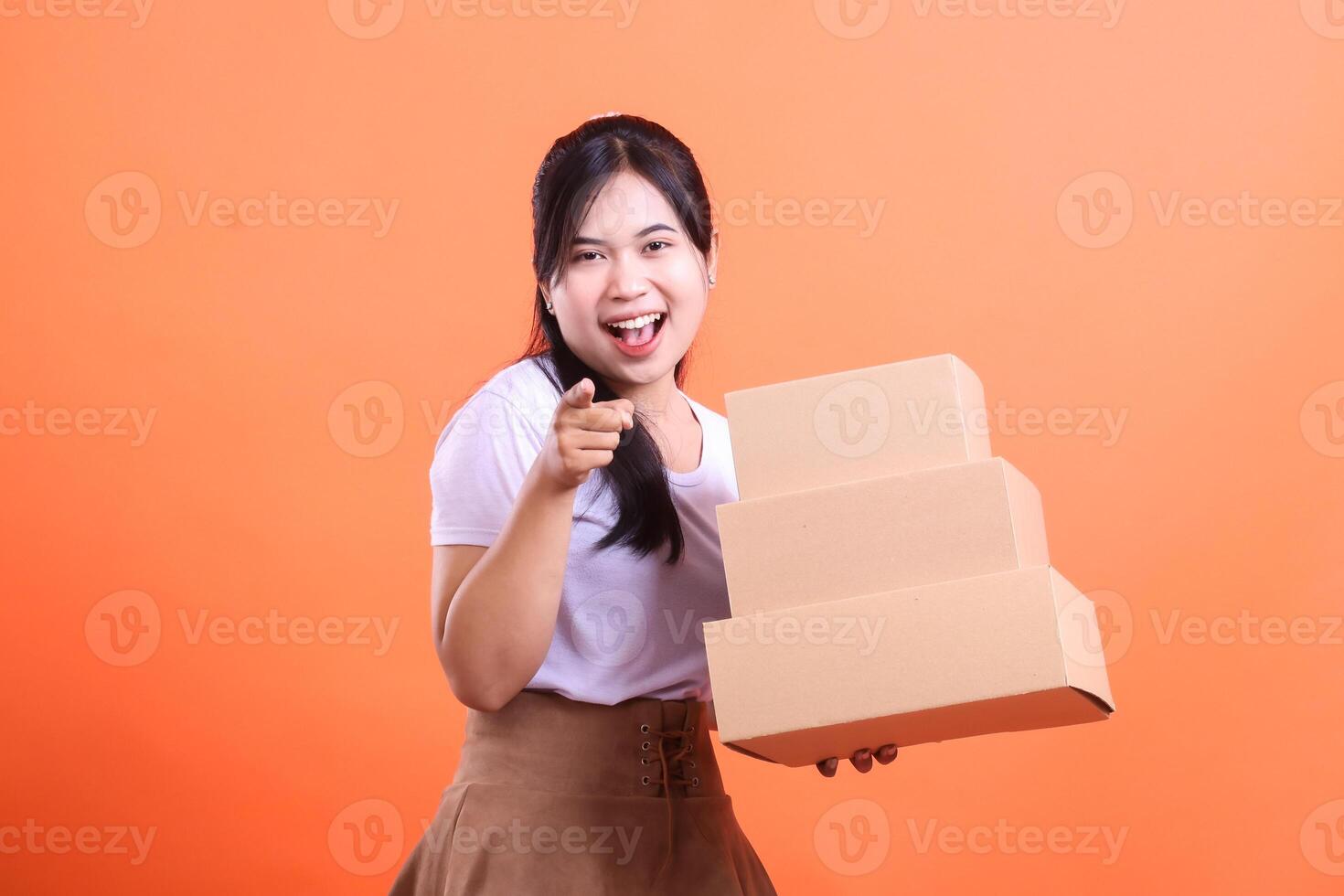 A young woman with a big smile is pointing at the camera while holding a stack of delivery boxes in her hands isolated on orange background. photo